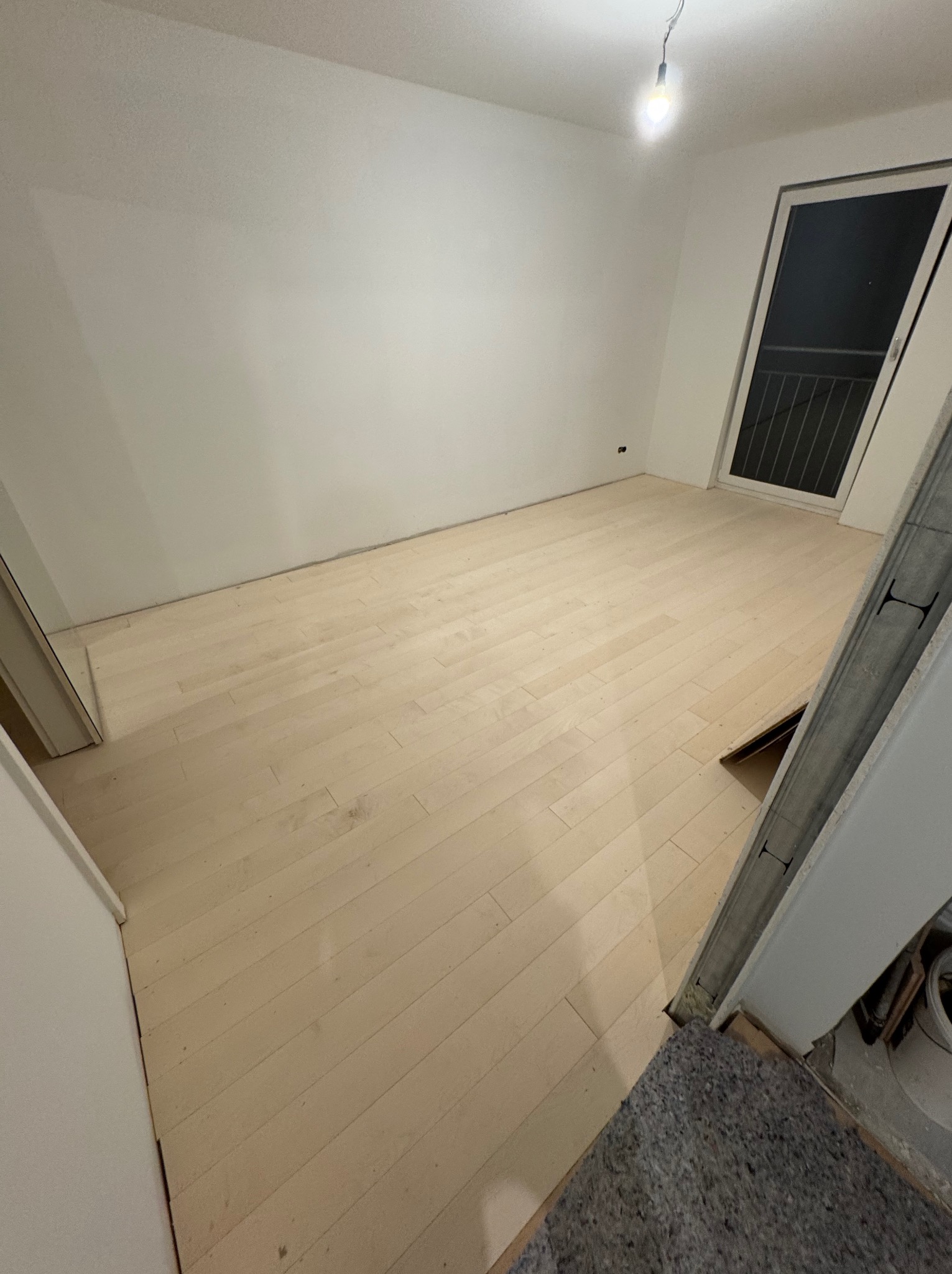 Jasne panele podłogowe w pustym, odświeżonym pokoju z białymi ścianami i oknem balkonowym. Widoczny fragment futryny drzwi i prowizoryczne oświetlenie.