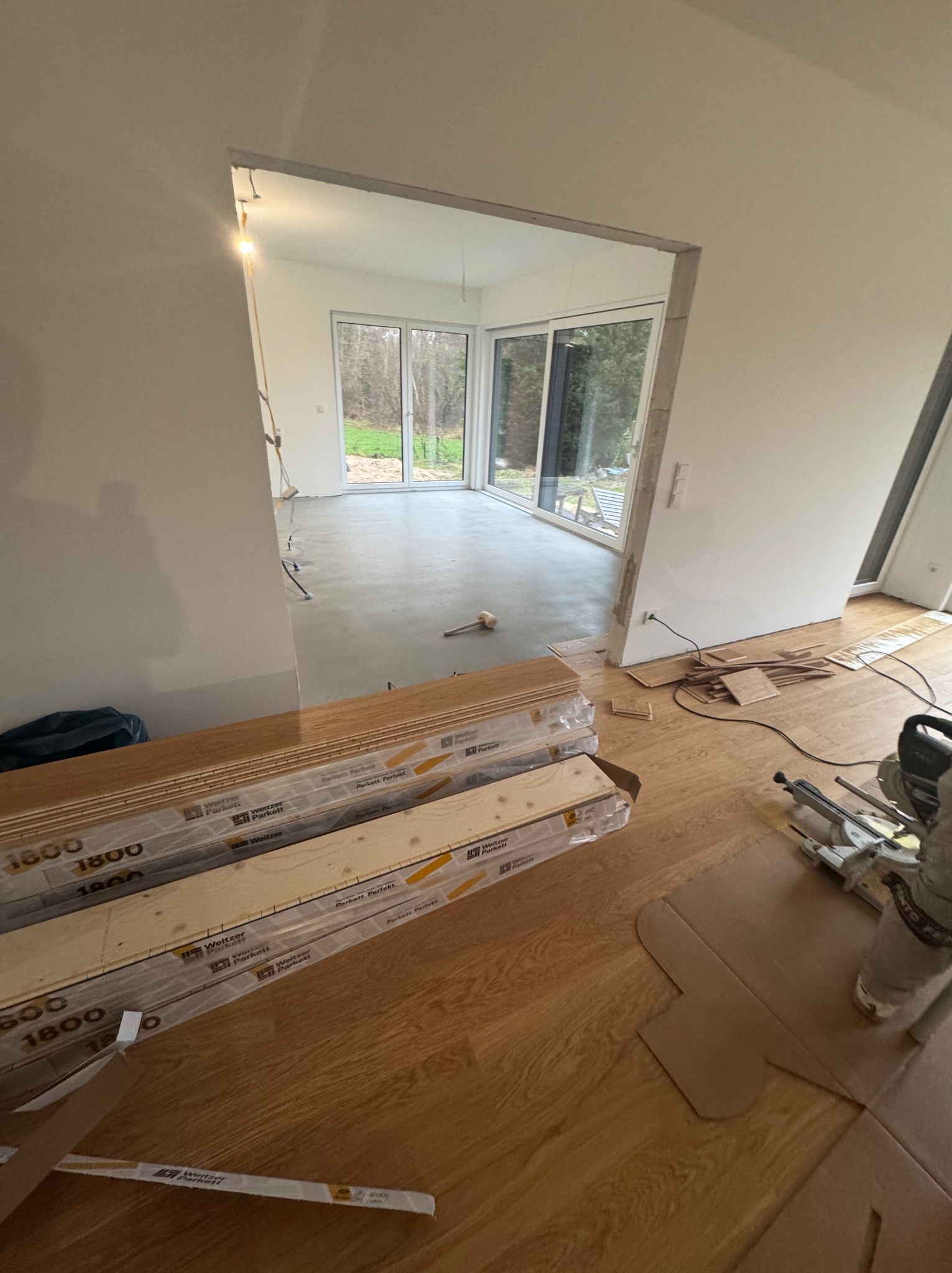 Układanie paneli: widok na pokój z częściowo ułożoną podłogą, paczki paneli Weitzer Parkett i piła ukośnica. W tle okna z widokiem na zieleń.