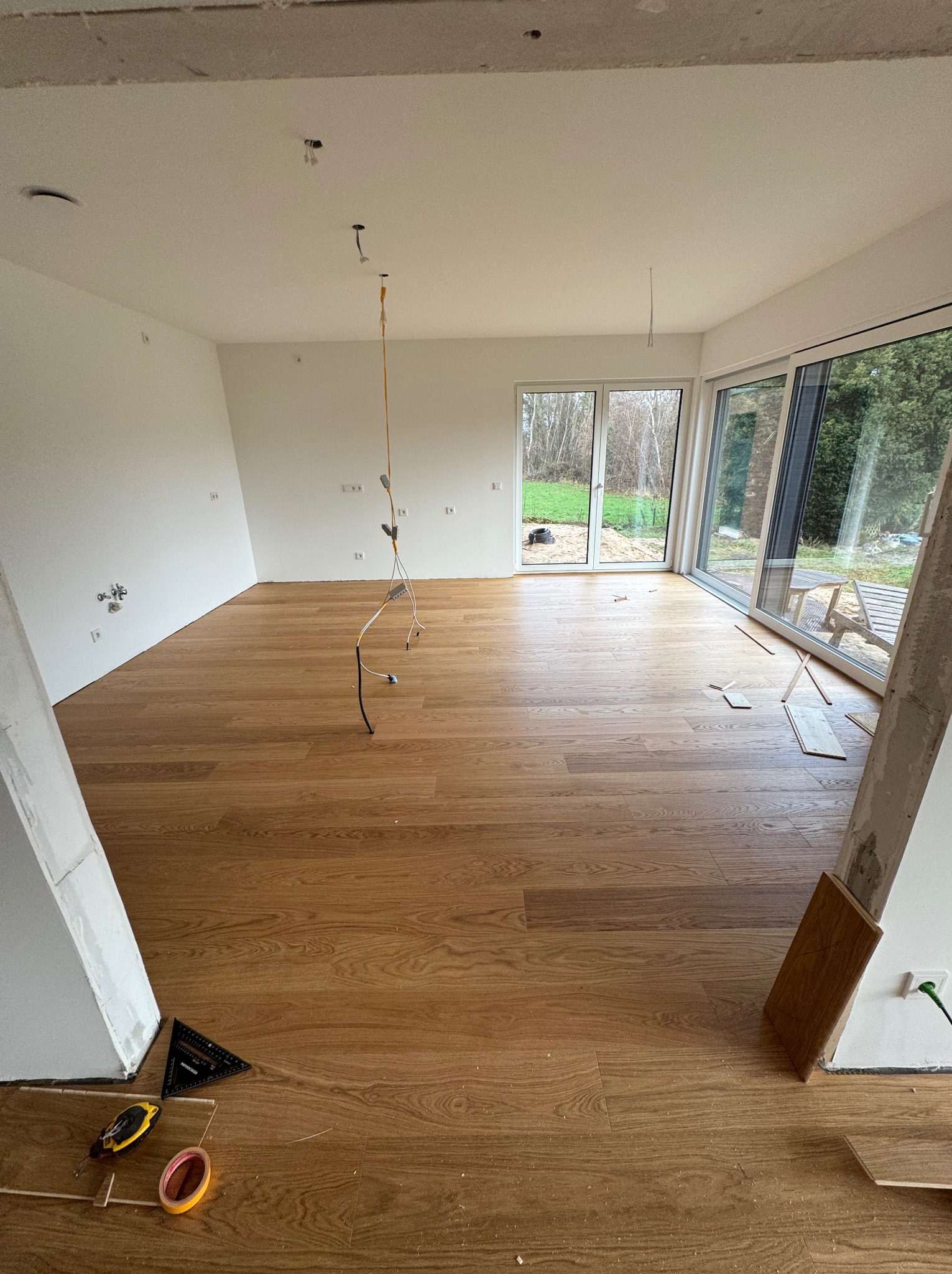 Jasne panele podłogowe ułożone w przestronnym pokoju z dużymi oknami. Widoczne narzędzia i niedokończone prace wykończeniowe przy ścianach. W tle zieleń za oknem.