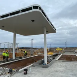 Budowa stacji paliw w trakcie realizacji: widoczne zbrojenia pod wylewkę, pracownicy w żółtych kamizelkach, stalowe konstrukcje w tle, pochmurne niebo.