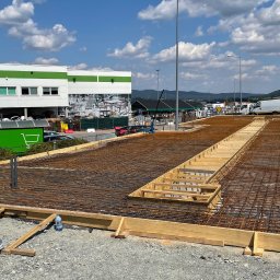 FA MATLAG MATLĄG STANISŁAW - Przygotowanie zbrojenia i szalunku pod fundamenty na placu budowy. Widoczne stalowe pręty, drewniane elementy konstrukcyjne oraz budynek w tle.
