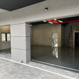 FA MATLAG MATLĄG STANISŁAW - Wnętrze budynku w surowym stanie z posadzką w kolorze grafitowym. Widoczne fragmenty ścian i sufitu. Płytki chodnikowe na zewnątrz budynku.