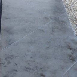 FA MATLAG MATLĄG STANISŁAW - Szara posadzka z mikrocementu na zewnątrz budynku, widoczne geometryczne podziały i odpływ liniowy. W tle mur z kamienia i elementy otoczenia.