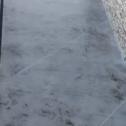 Szara posadzka z mikrocementu na zewnątrz budynku, widoczne geometryczne podziały i odpływ liniowy. W tle mur z kamienia i elementy otoczenia.