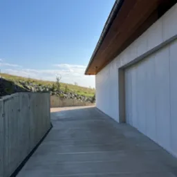 Nowoczesny budynek z białymi panelami elewacyjnymi i betonową drogą dojazdową. Widoczny fragment muru oporowego i wzgórze w tle.