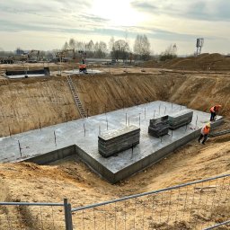 FA MATLAG MATLĄG STANISŁAW - Betonowy fundament w wykopie, widoczni pracownicy w odblaskowych kamizelkach. Na placu budowy sprzęt i materiały budowlane, zabezpieczenia. Ujęcie z góry.