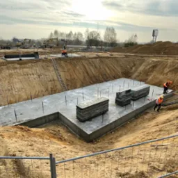 Betonowy fundament w wykopie, widoczni pracownicy w odblaskowych kamizelkach. Na placu budowy sprzęt i materiały budowlane, zabezpieczenia. Ujęcie z góry.