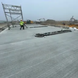 Świeżo wylana, gładka powierzchnia betonowa z wycięciami na placu budowy w mglisty dzień. W tle sprzęt budowlany i pracownicy w odblaskowych kamizelkach.