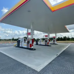 Stacja paliw Shell z dystrybutorami V-Power, widok z perspektywy posadzki z kostki brukowej przechodzącej w betonową wylewkę pod zadaszeniem.