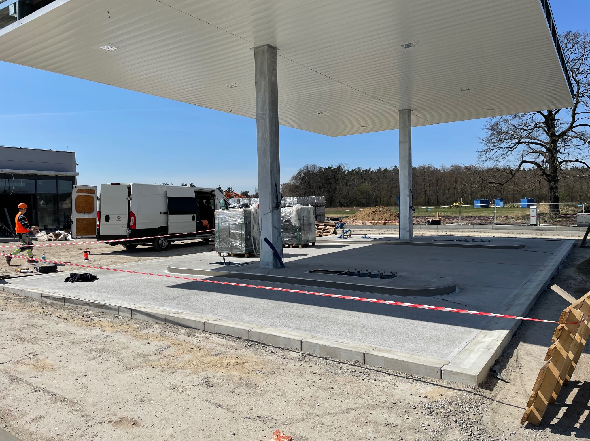 Nowa, betonowa posadzka stacji paliw z obrzeżami, zabezpieczona taśmą ostrzegawczą. W tle biały van i pracownik w odblaskowej kamizelce. Słoneczny dzień.