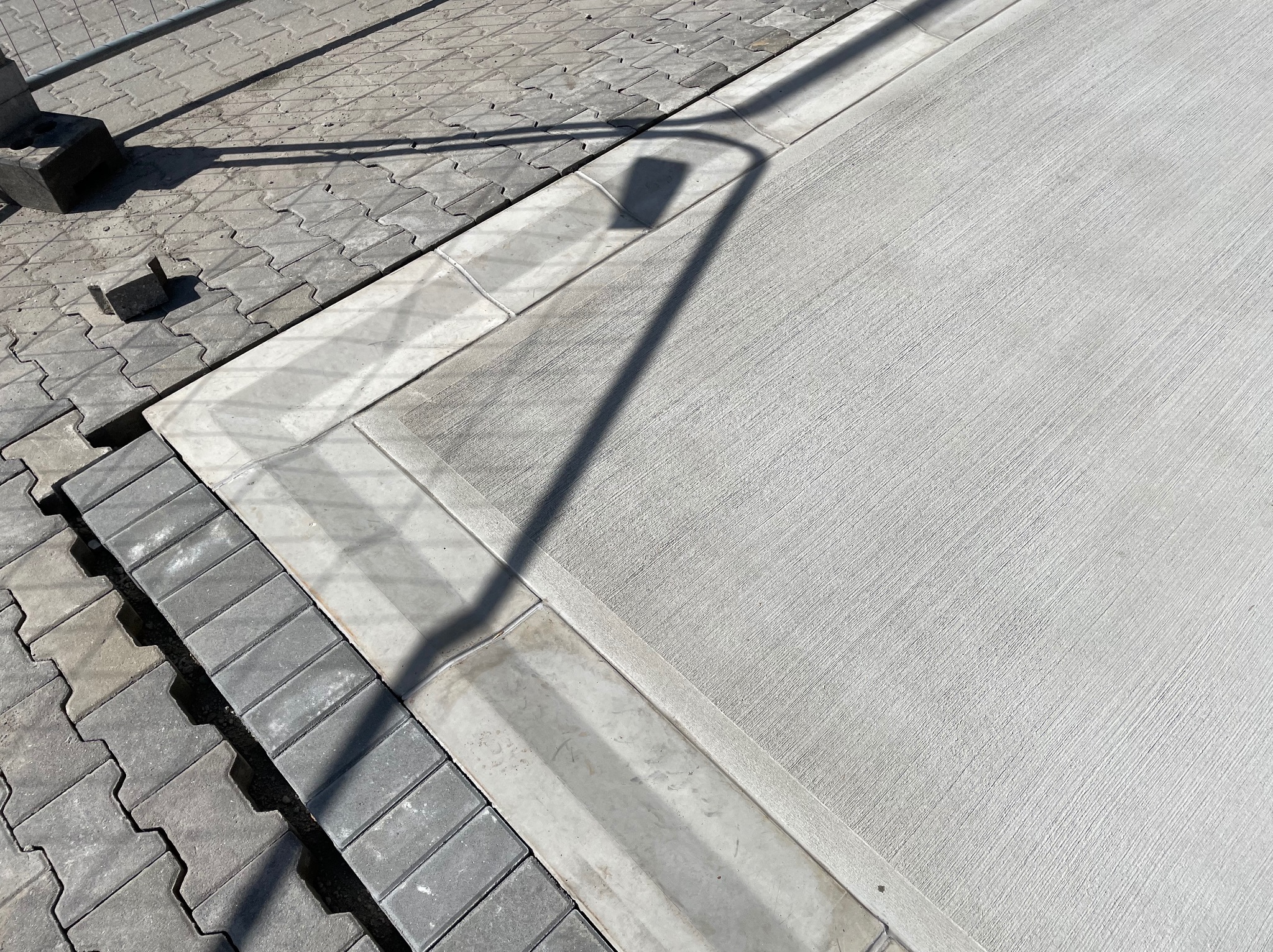 Świeżo wylana, jasnoszara wylewka betonowa granicząca z szarą kostką brukową. Widoczny cień lampy ulicznej. Detal wykończenia powierzchni.