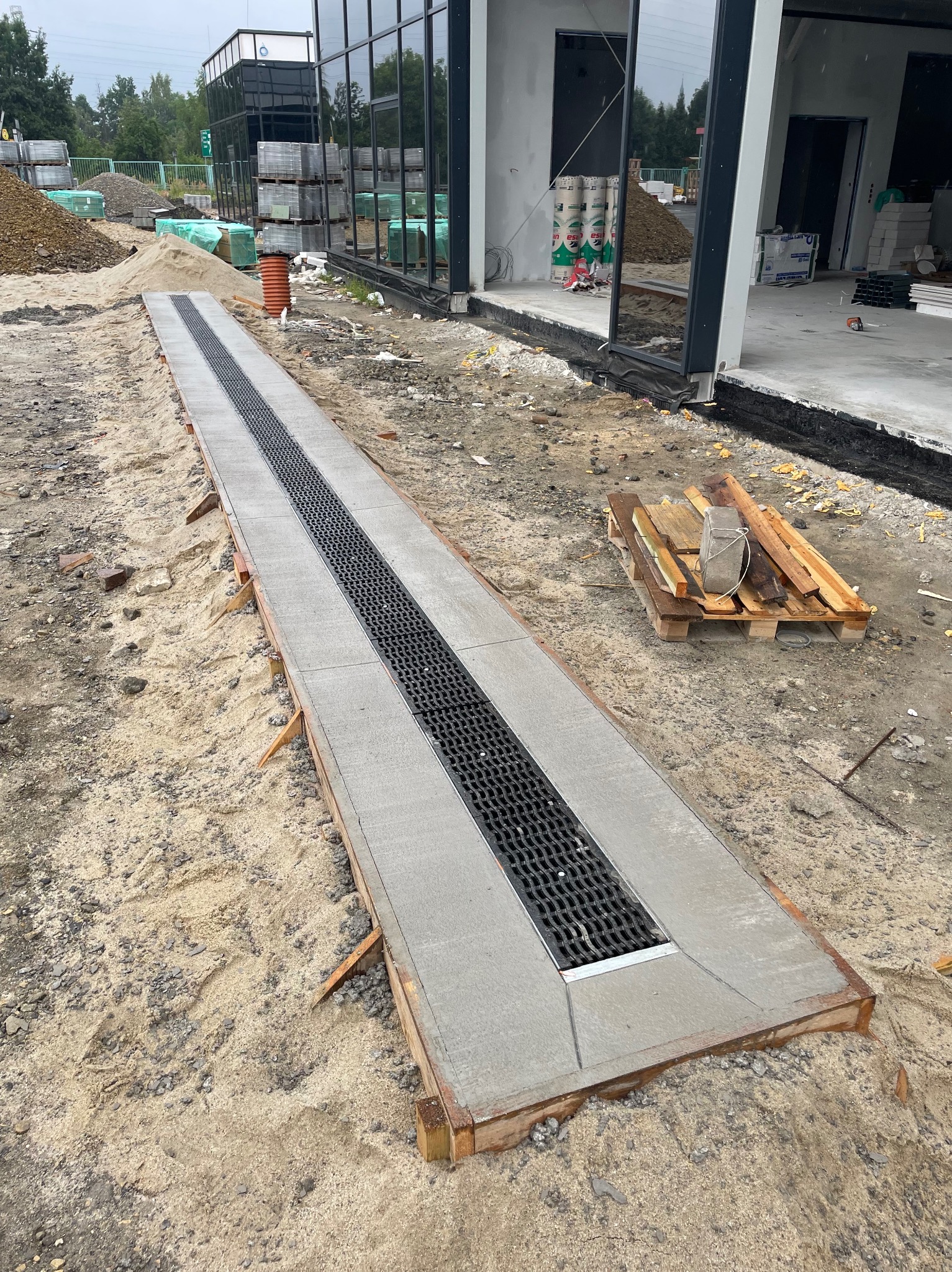 Długi, betonowy odpływ liniowy z rusztem, zamontowany przed budynkiem w trakcie prac wykończeniowych. Piasek i materiały budowlane w tle.