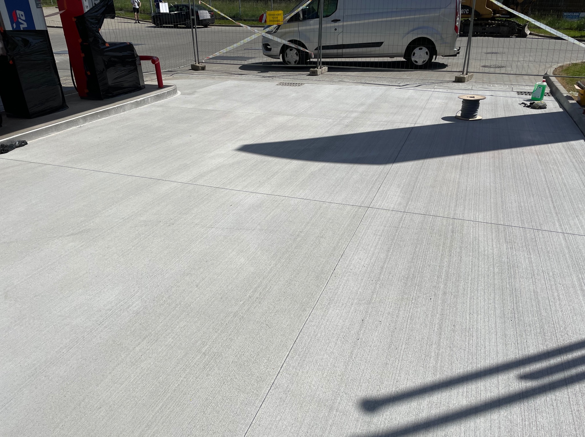 Świeżo wykonana, szara posadzka przemysłowa z widocznymi nacięciami dylatacyjnymi. W tle dystrybutory paliw i zaparkowany bus.