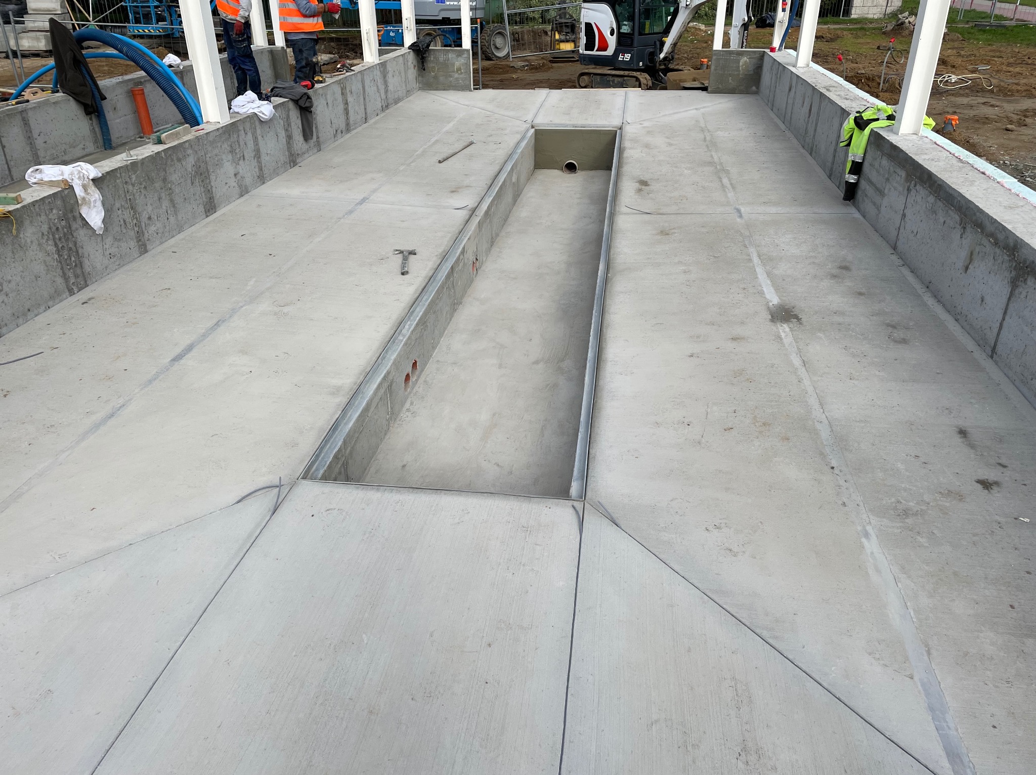 Szara, betonowa konstrukcja z zagłębieniem w centralnej części, widoczna z góry. Elementy budowlane, sprzęt i pracownicy w tle. Widoczne ślady po szalunku.