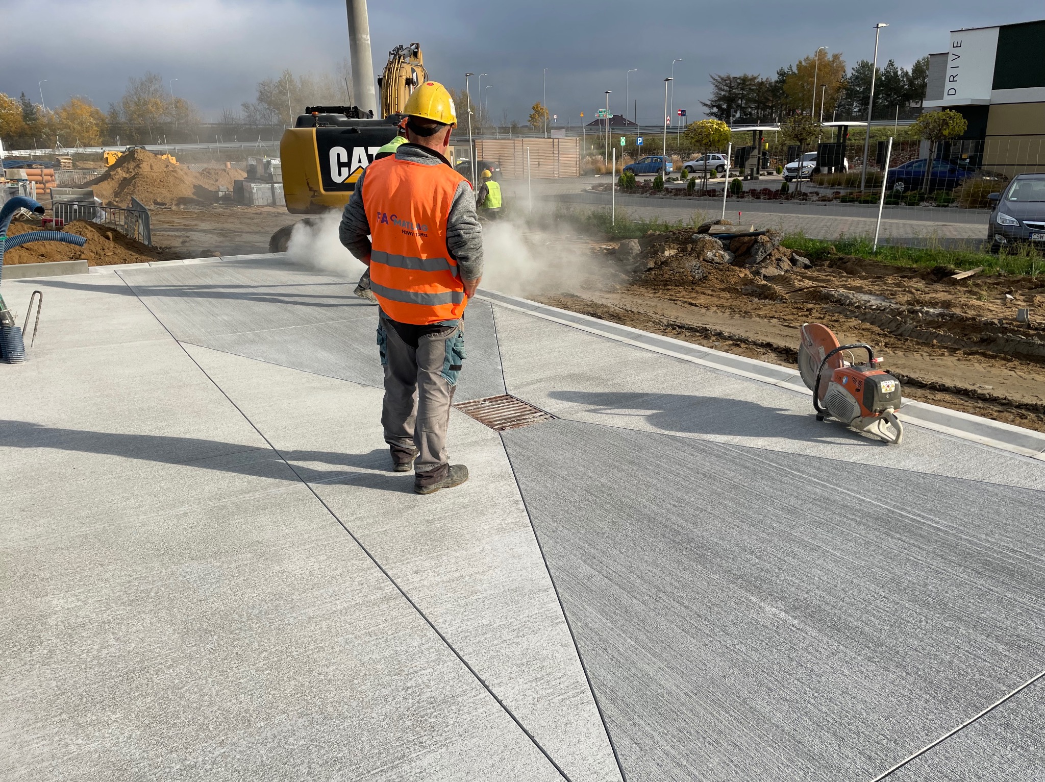Pracownik w kasku i kamizelce na tle świeżo wykonanej posadzki przemysłowej z widocznymi liniami cięcia dylatacyjnego i maszyną do cięcia. W tle koparka.