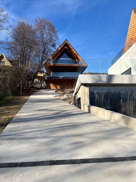 Nowoczesny dom z drewnianą elewacją i szklanymi balustradami, widziany z perspektywy betonowego chodnika w słoneczny dzień. Unikalna bryła budynku.