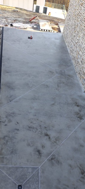 Szara posadzka z mikrocementu na zewnątrz budynku, widoczne geometryczne podziały i odpływ liniowy. W tle mur z kamienia i elementy otoczenia.