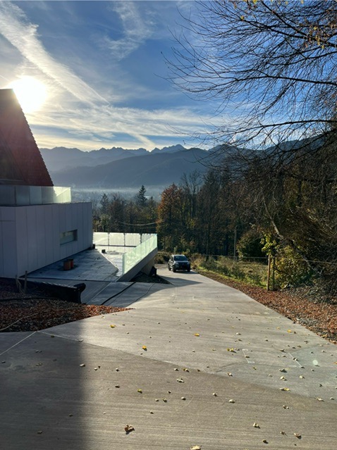 Nowoczesny dom z betonowym podjazdem na tle gór. Widoczny fragment budynku z przeszkloną balustradą i samochód na podjeździe. Jesienne liście na betonowej powierzchni.