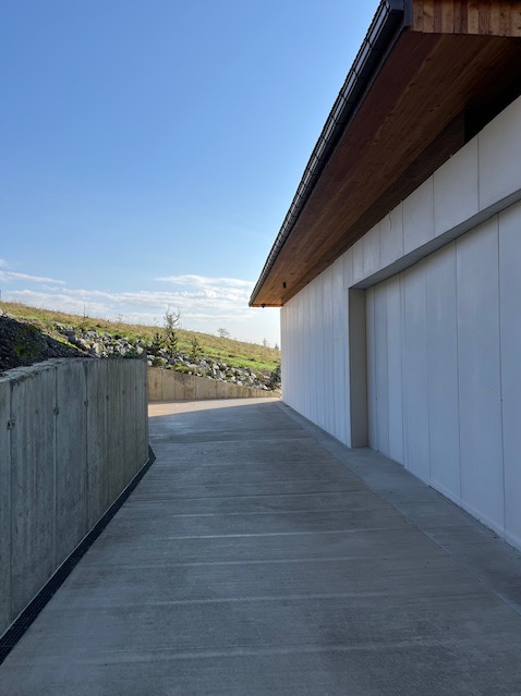 Nowoczesny budynek z białymi panelami elewacyjnymi i betonową drogą dojazdową. Widoczny fragment muru oporowego i wzgórze w tle.