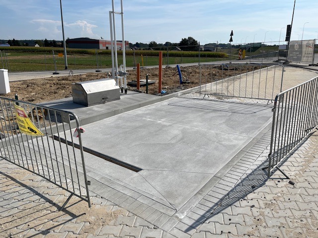 Świeża, gładka powierzchnia betonowa z odpływem liniowym, zabezpieczona barierkami na placu budowy. Widoczna metalowa skrzynka i instalacje. Słoneczny dzień.