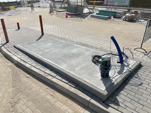 Wykończona, prostokątna platforma z szarego betonu otoczona obrzeżem, na tle kostki brukowej i elementów placu budowy. Widoczne kable i rury.