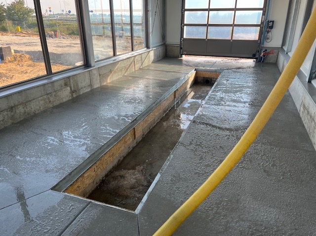 Świeżo wylana, mokra posadzka w warsztacie samochodowym z kanałem rewizyjnym. W tle brama garażowa i okna z widokiem na plac budowy.