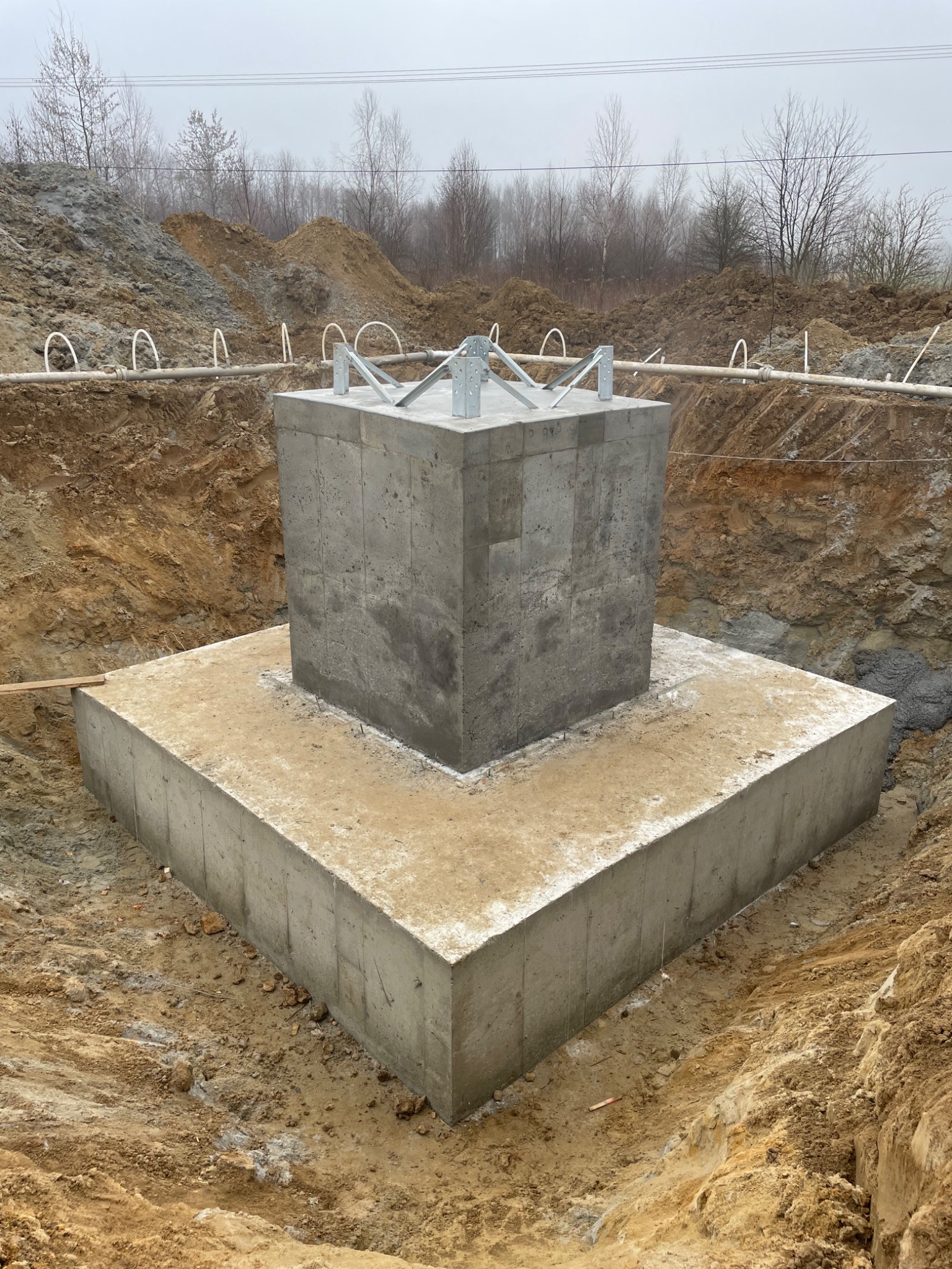 Żelbetowy fundament pod budowę w Nowym Targu. Widoczna podstawa i pionowy słup. Wokół wykop, ziemia i elementy zbrojenia. Pochmurny dzień.
