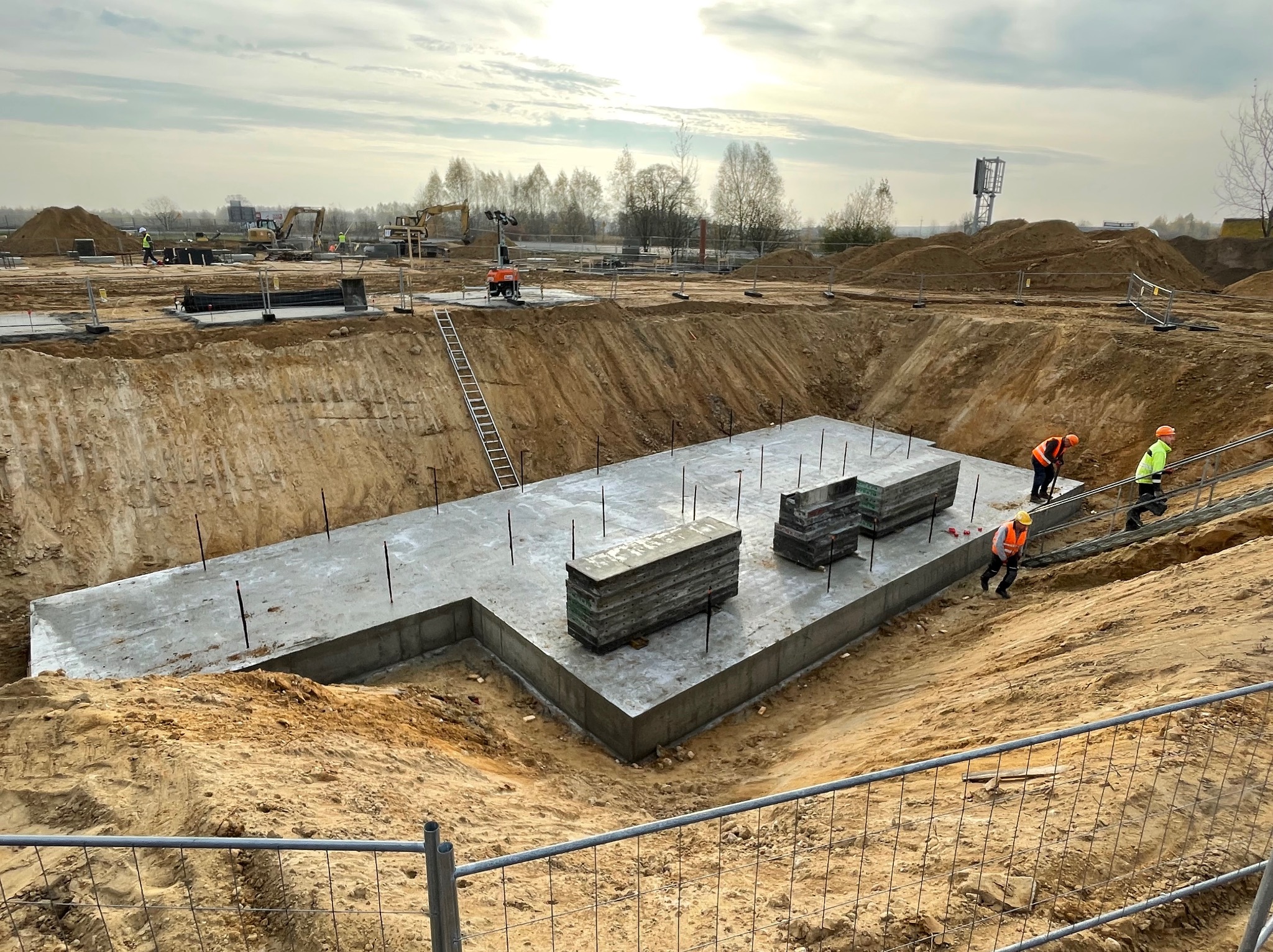 Betonowy fundament w wykopie, widoczni pracownicy w odblaskowych kamizelkach. Na placu budowy sprzęt i materiały budowlane, zabezpieczenia. Ujęcie z góry.
