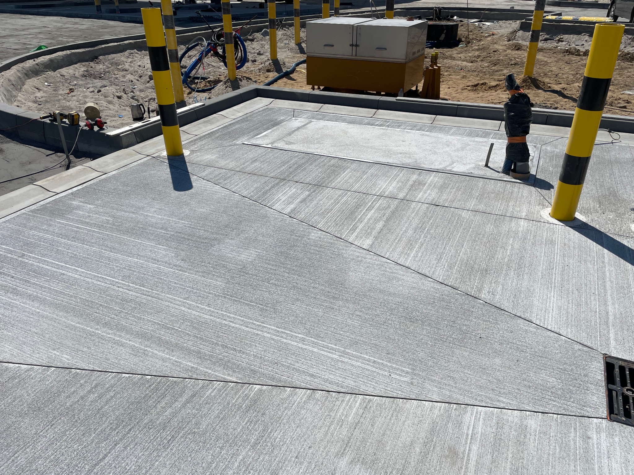 Szara, gładka powierzchnia z betonu podzielona na segmenty, z widocznym odpływem i słupkami ostrzegawczymi w tle. Prace wykończeniowe w przestrzeni publicznej.