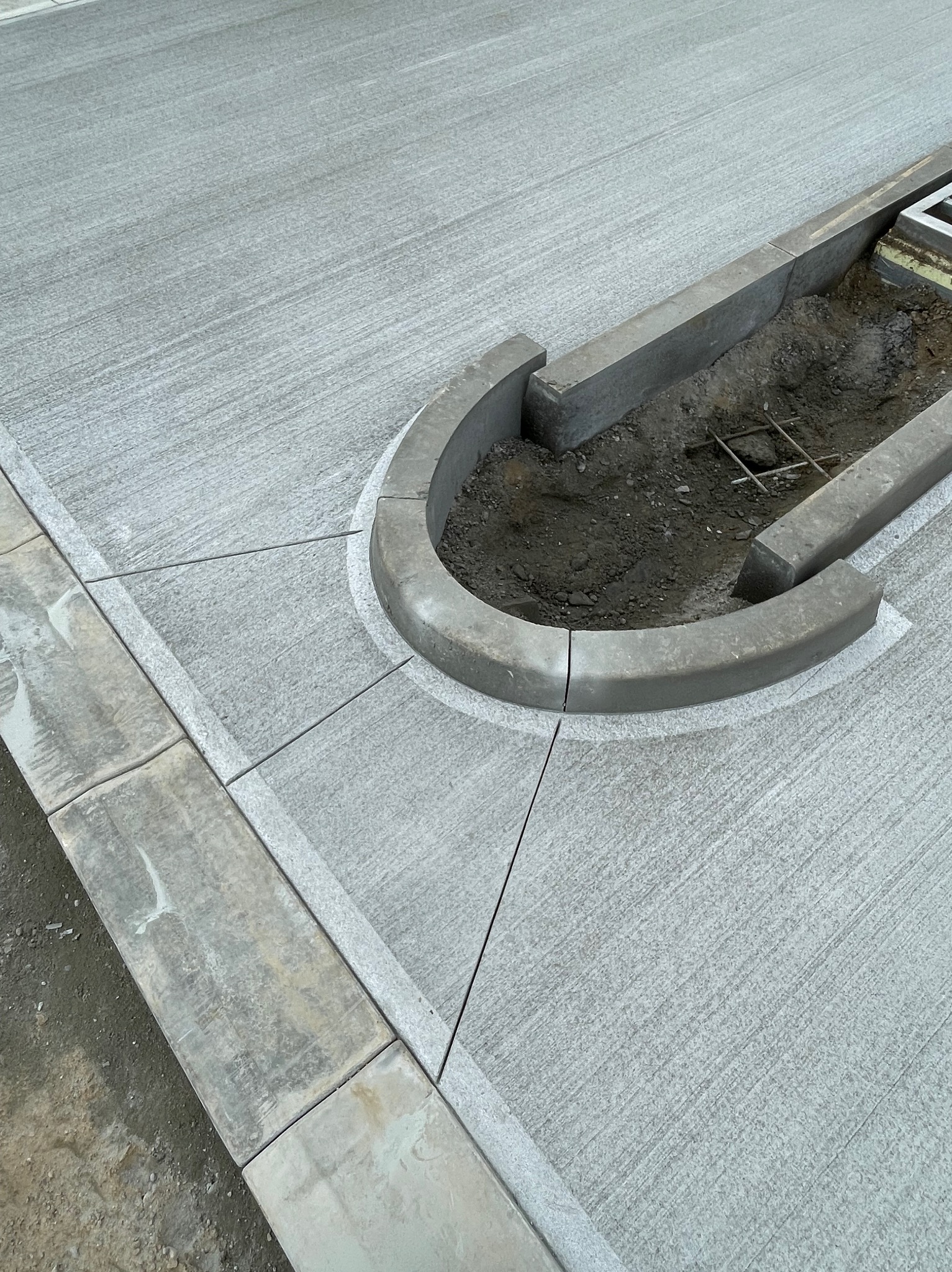 Szara, teksturowana nawierzchnia betonowa z obrzeżami i krawężnikami. Widoczny fragment studzienki kanalizacyjnej i zbrojenie w wykopie.