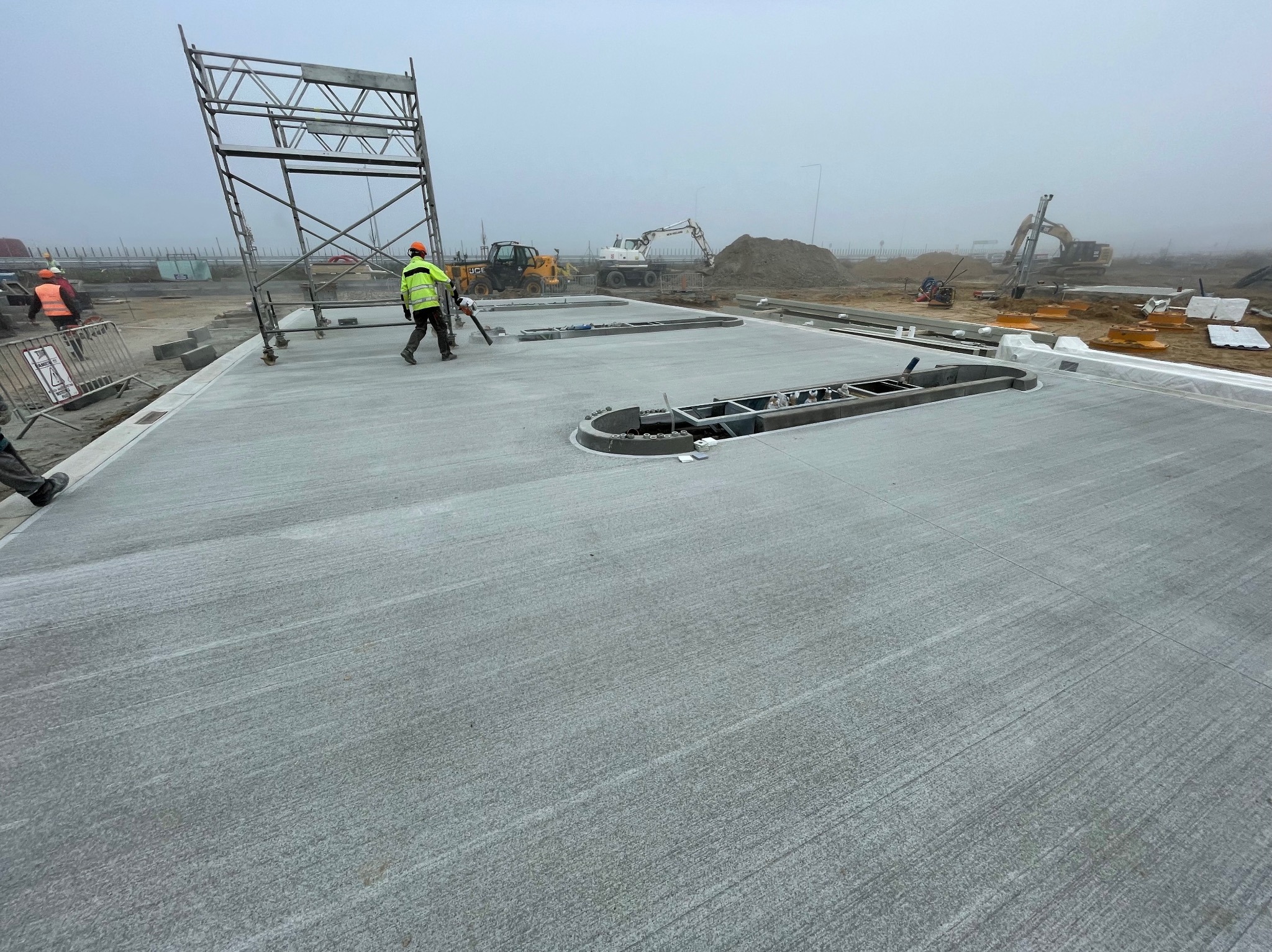 Świeżo wylana, gładka powierzchnia betonowa z wycięciami na placu budowy w mglisty dzień. W tle sprzęt budowlany i pracownicy w odblaskowych kamizelkach.