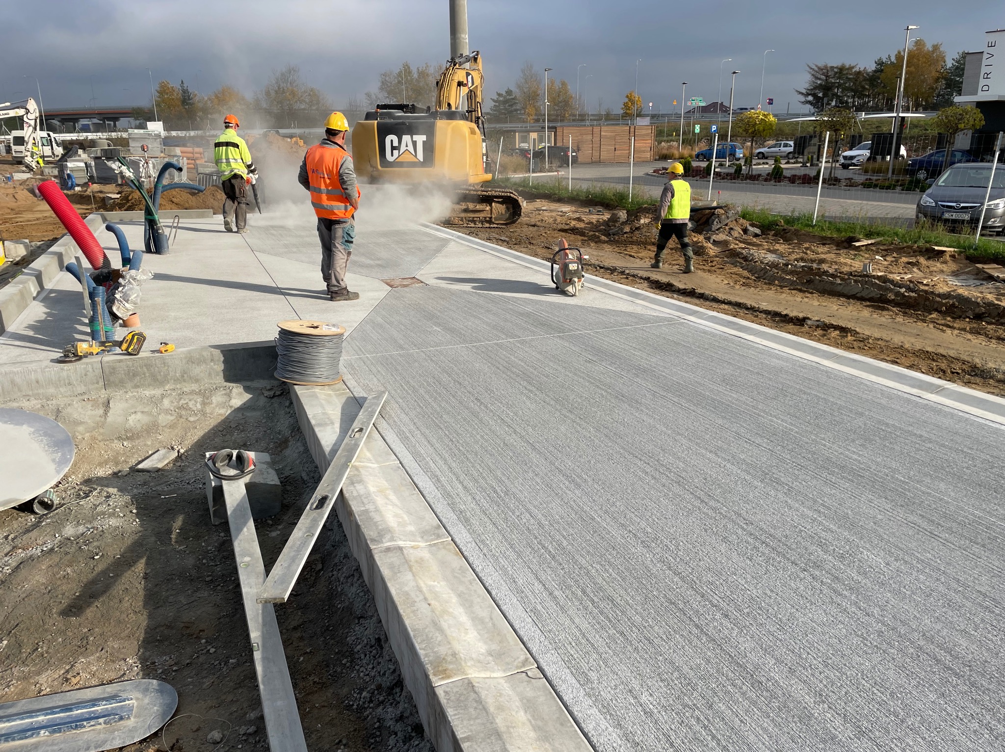 Świeżo wylana betonowa nawierzchnia z pracownikami i sprzętem budowlanym w tle, w tym koparka CAT. Widoczne narzędzia i akcesoria do obróbki betonu.