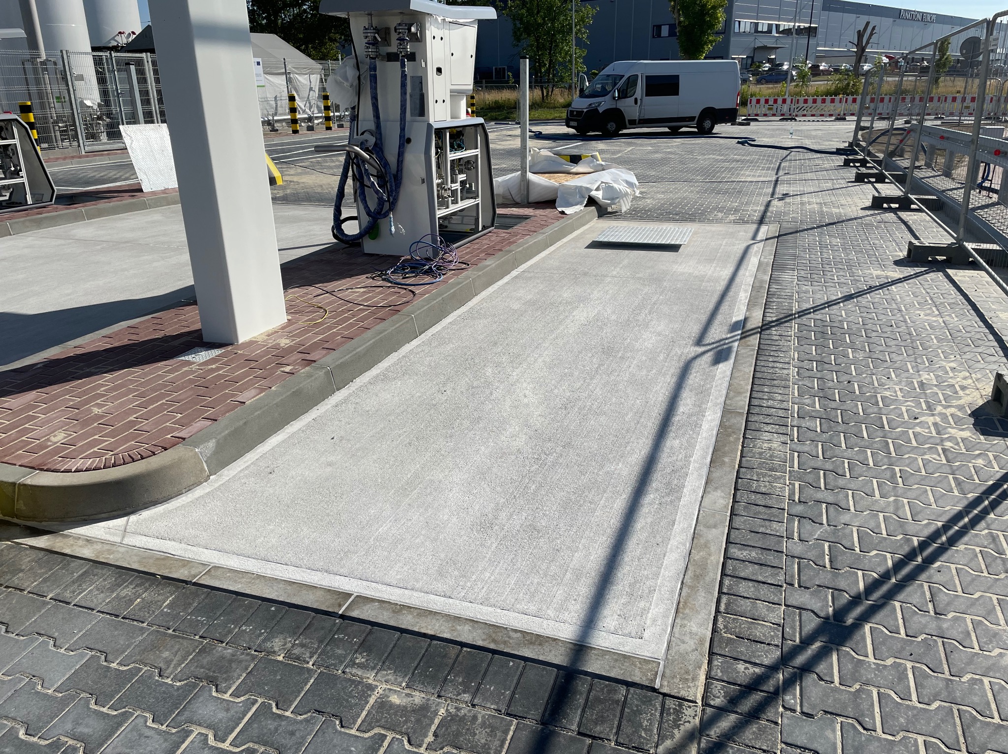 Nowa, jasnoszara posadzka z betonu z wykończeniem obrzeżem, otoczona kostką brukową. W tle biały samochód dostawczy i budynek przemysłowy Panattoni Europe.