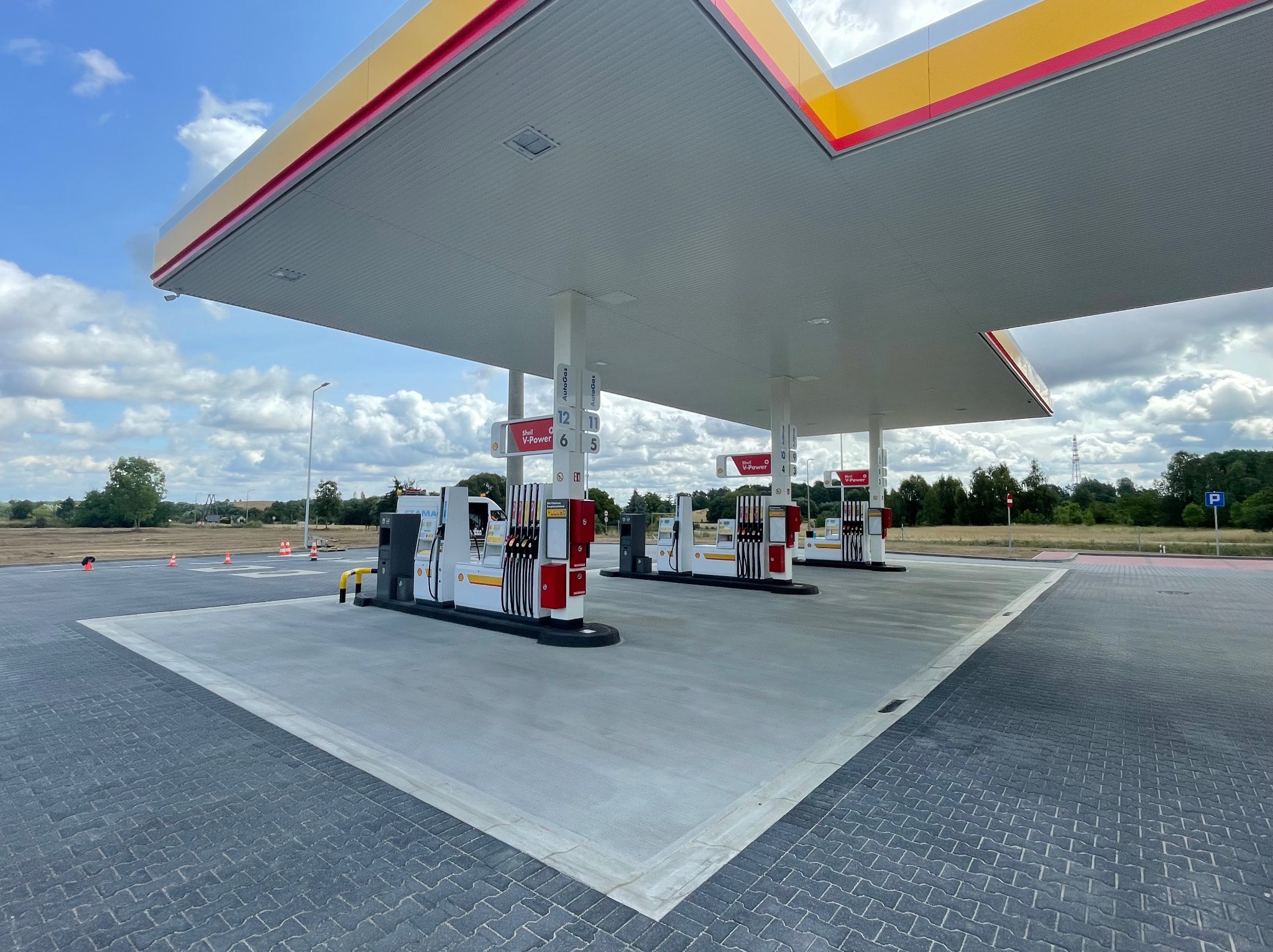 Stacja paliw Shell z dystrybutorami V-Power, widok z perspektywy posadzki z kostki brukowej przechodzącej w betonową wylewkę pod zadaszeniem.