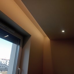 Paweł Żoch - Pomalowane na beżowo ściany i sufit w rogu pokoju z oknem, widok na plac budowy za oknem, oświetlenie punktowe w suficie.