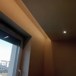 Pomalowane na beżowo ściany i sufit w rogu pokoju z oknem, widok na plac budowy za oknem, oświetlenie punktowe w suficie.