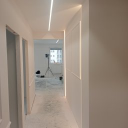 Paweł Żoch - Korytarz w trakcie prac wykończeniowych, podłoga zabezpieczona folią malarską, widoczne oświetlenie LED w listwach oraz lampa na statywie w pokoju obok.