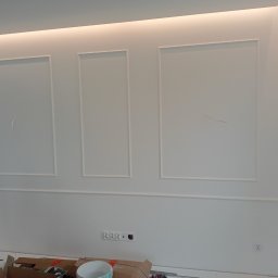 Paweł Żoch - Biała ściana z dekoracyjnymi listwami w kształcie prostokątów, widoczne gniazdka elektryczne i elementy robocze na podłodze, oświetlenie LED na suficie.