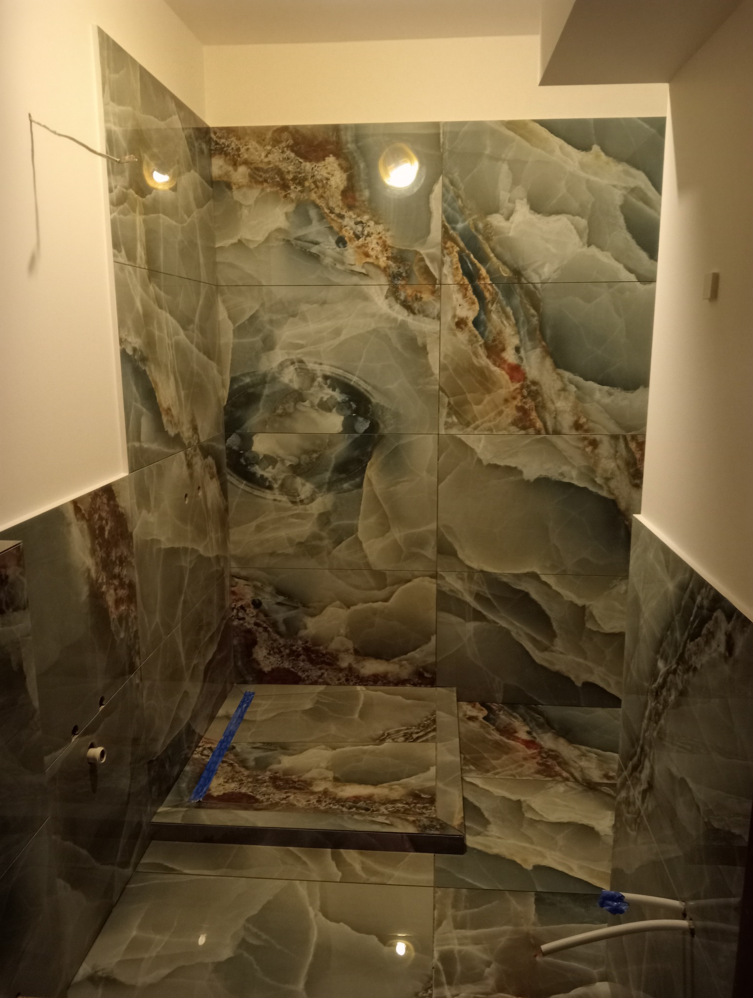 Wnętrze łazienki w trakcie wykańczania, widoczne płytki imitujące marmur na ścianach i podłodze, wystające rury instalacji wodnej oraz taśma malarska zabezpieczająca krawędzie.