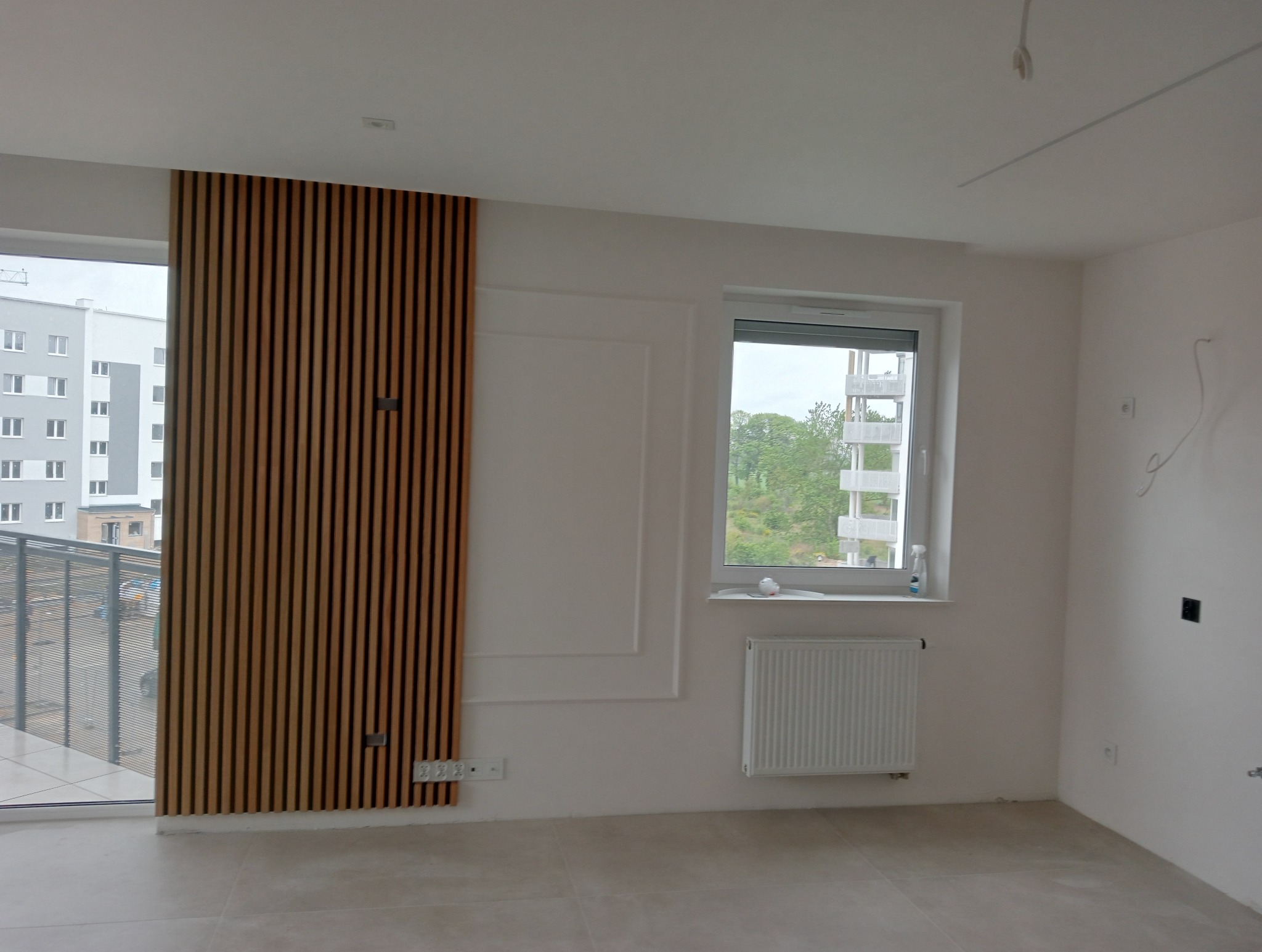 Nowoczesne wykończenie wnętrza: drewniane lamele na ścianie obok okna balkonowego, białe ściany z dekoracyjnymi panelami, grzejnik pod oknem, widok na szary budynek za oknem.