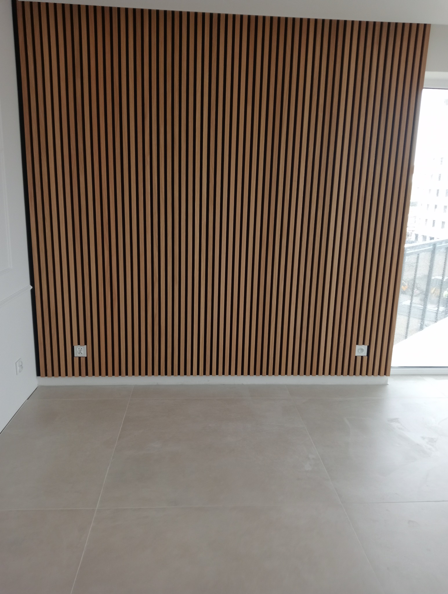 Ściana z drewnianych paneli akustycznych w pionowym układzie, widoczne gniazdka elektryczne, jasna podłoga z dużych płytek.