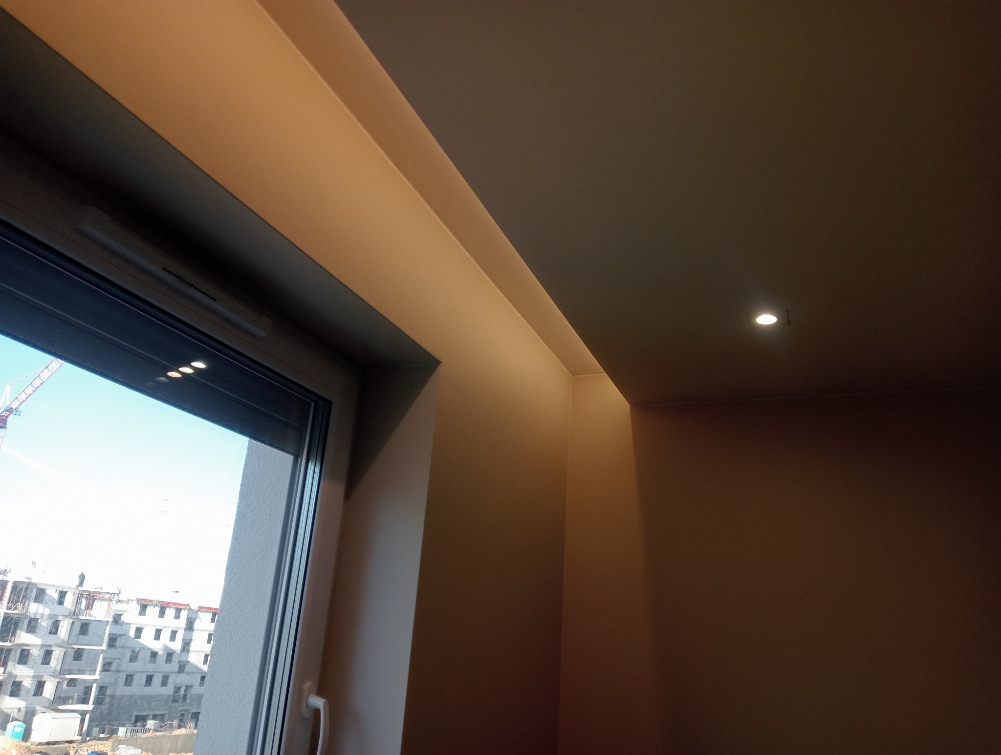 Pomalowane na beżowo ściany i sufit w rogu pokoju z oknem, widok na plac budowy za oknem, oświetlenie punktowe w suficie.