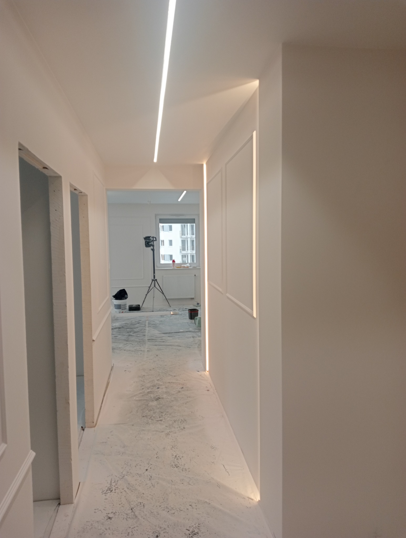 Korytarz w trakcie prac wykończeniowych, podłoga zabezpieczona folią malarską, widoczne oświetlenie LED w listwach oraz lampa na statywie w pokoju obok.