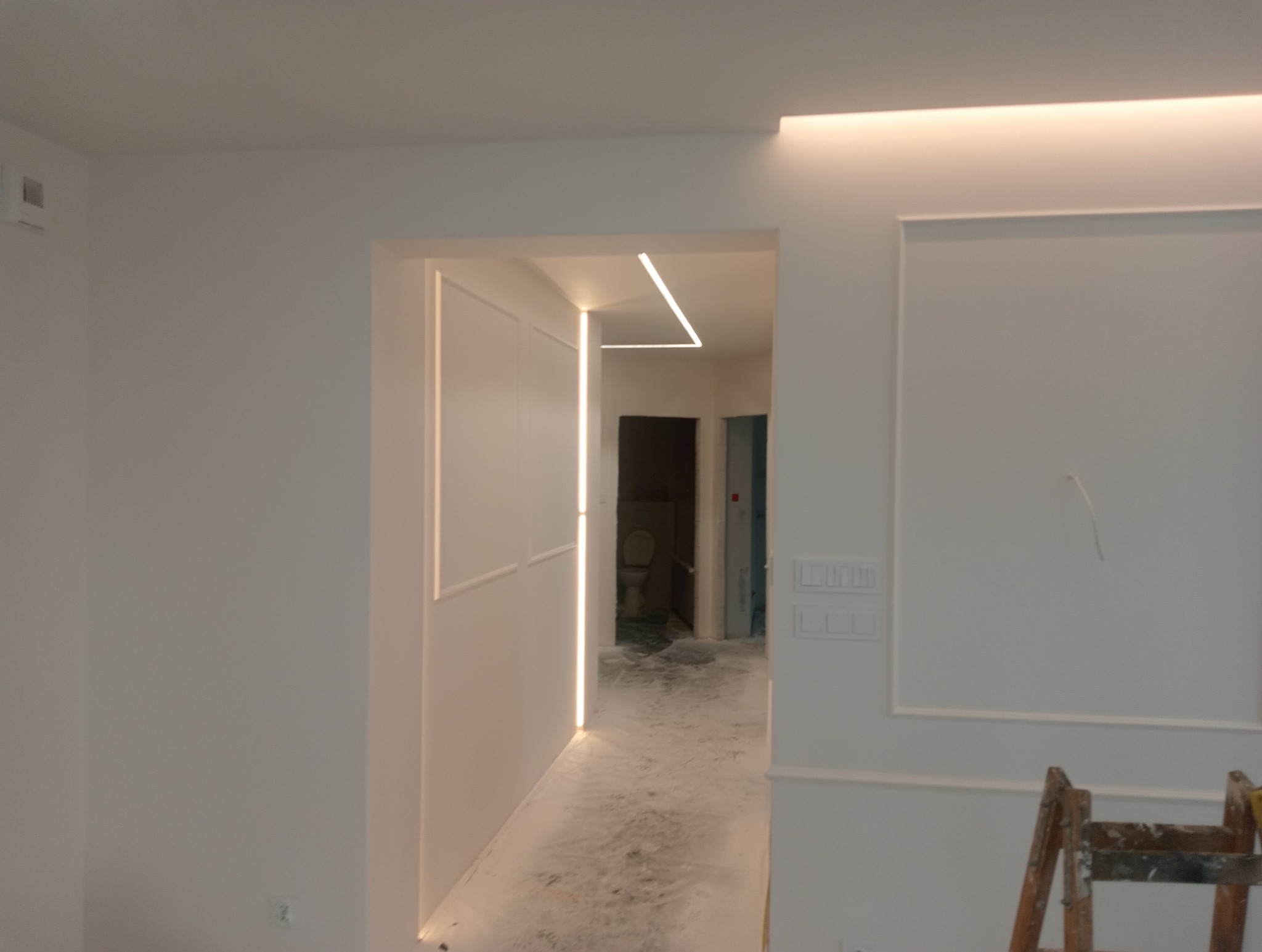 Nowoczesne wnętrze w trakcie prac wykończeniowych: białe ściany z dekoracyjnymi listwami, oświetlenie LED w suficie i wnękach, widoczna drabina malarska i częściowo zabezpieczona podłoga.