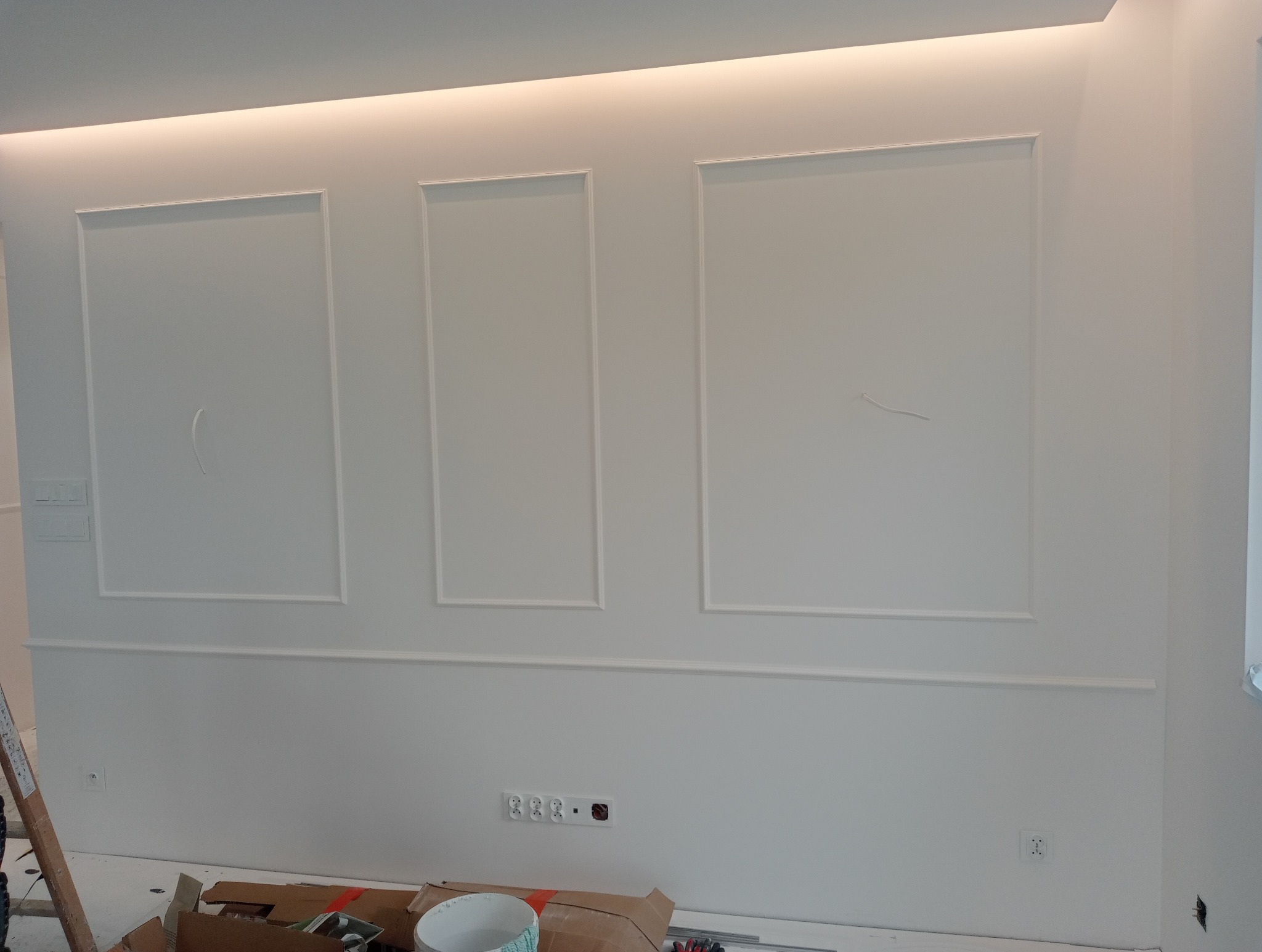 Biała ściana z dekoracyjnymi listwami w kształcie prostokątów, widoczne gniazdka elektryczne i elementy robocze na podłodze, oświetlenie LED na suficie.