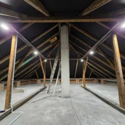 Poddasze z widoczną konstrukcją drewnianą dachu, oświetleniem LED zamontowanym na belkach i betonową podłogą. Widoczna drabina oparta o centralny filar.