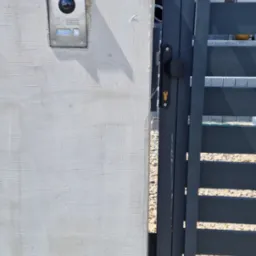 Zamontowany wideodomofon Hikvision na surowym, jasnym murze przy wejściowej furtce z metalowymi, poziomymi szczeblami.