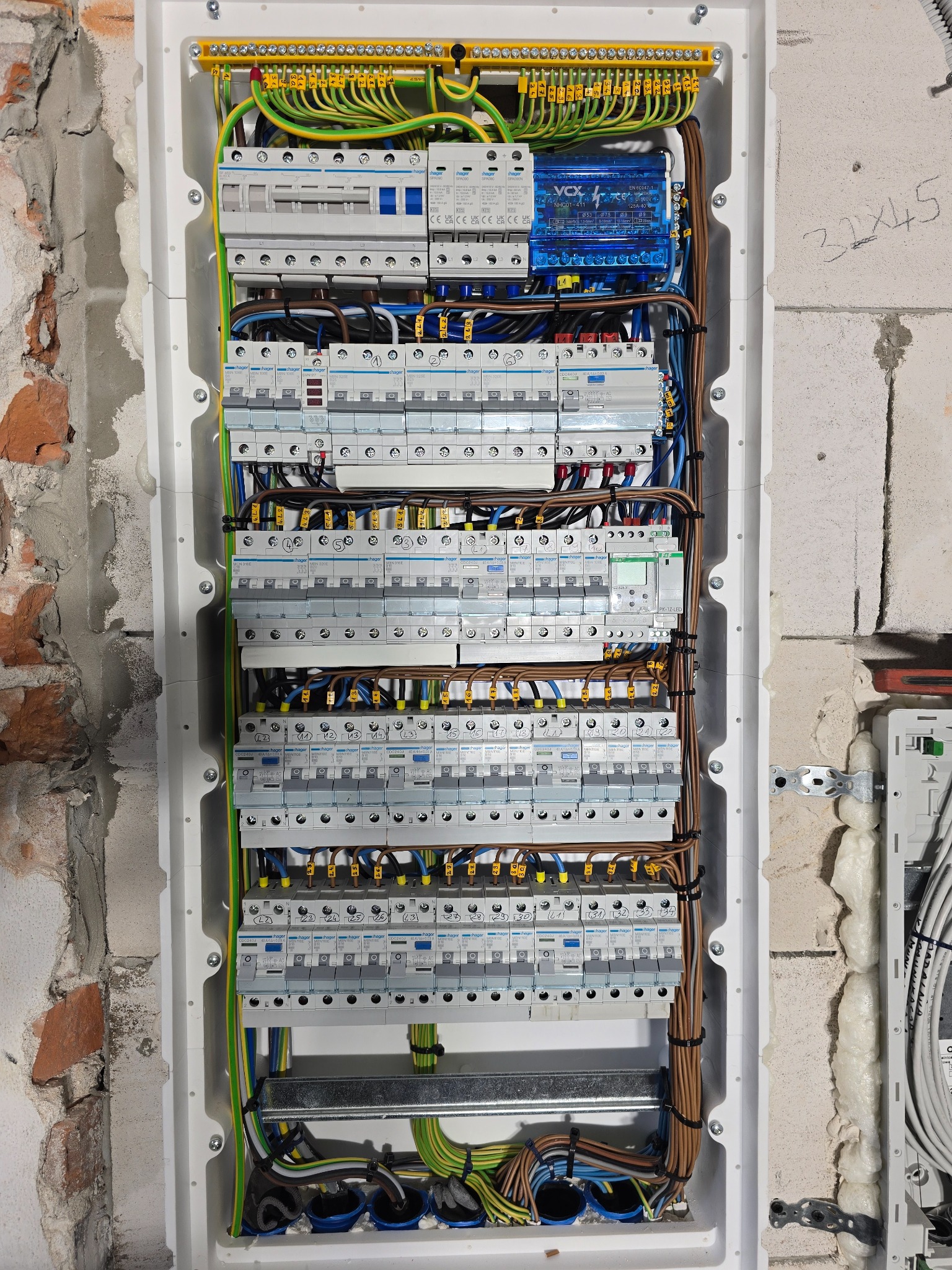 Rozdzielnica elektryczna z zamontowanymi bezpiecznikami i okablowaniem, widoczna na tle surowej ściany z cegły i betonu, z numeracją przewodów na żółtych etykietach.