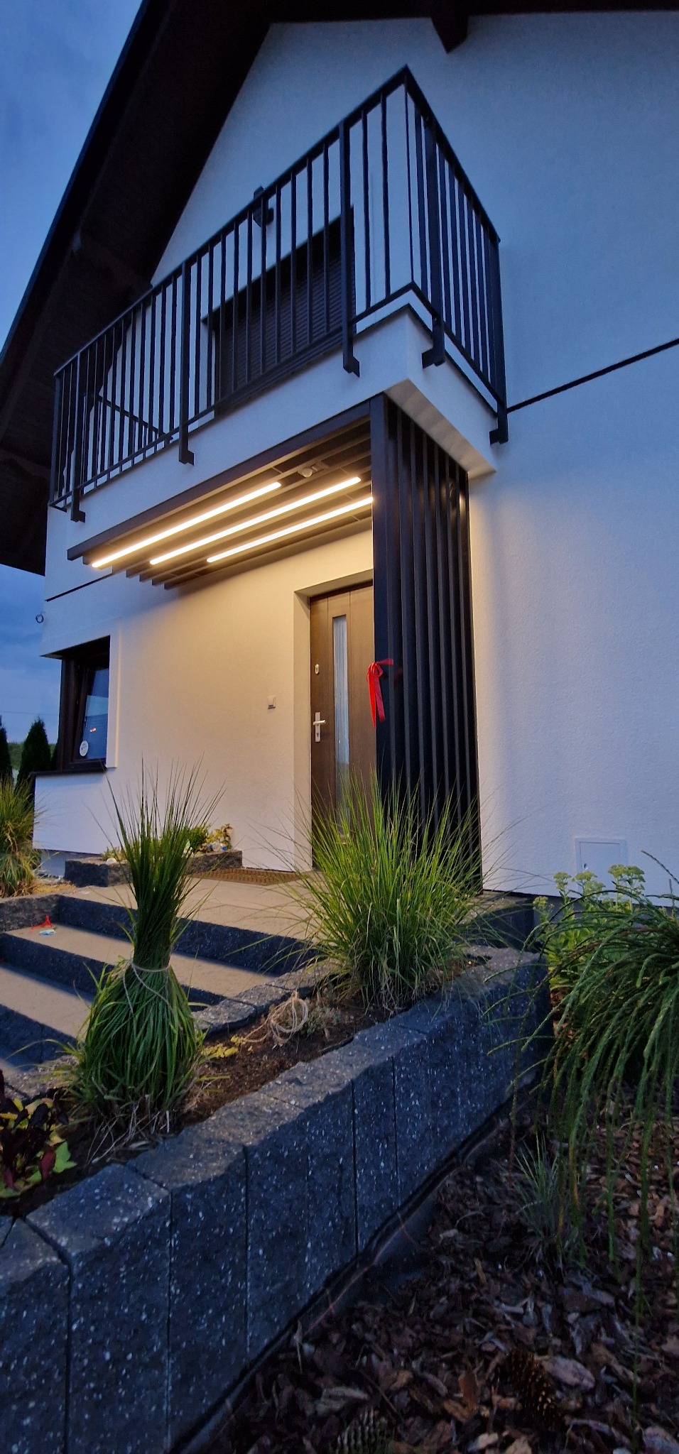 Nowoczesny dom z podświetlonym wejściem i balkonem, widok z dołu na elewację z elementami dekoracyjnymi i roślinnością.