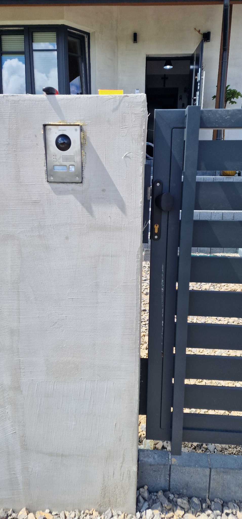 Zamontowany wideodomofon Hikvision na surowym, jasnym murze przy wejściowej furtce z metalowymi, poziomymi szczeblami.